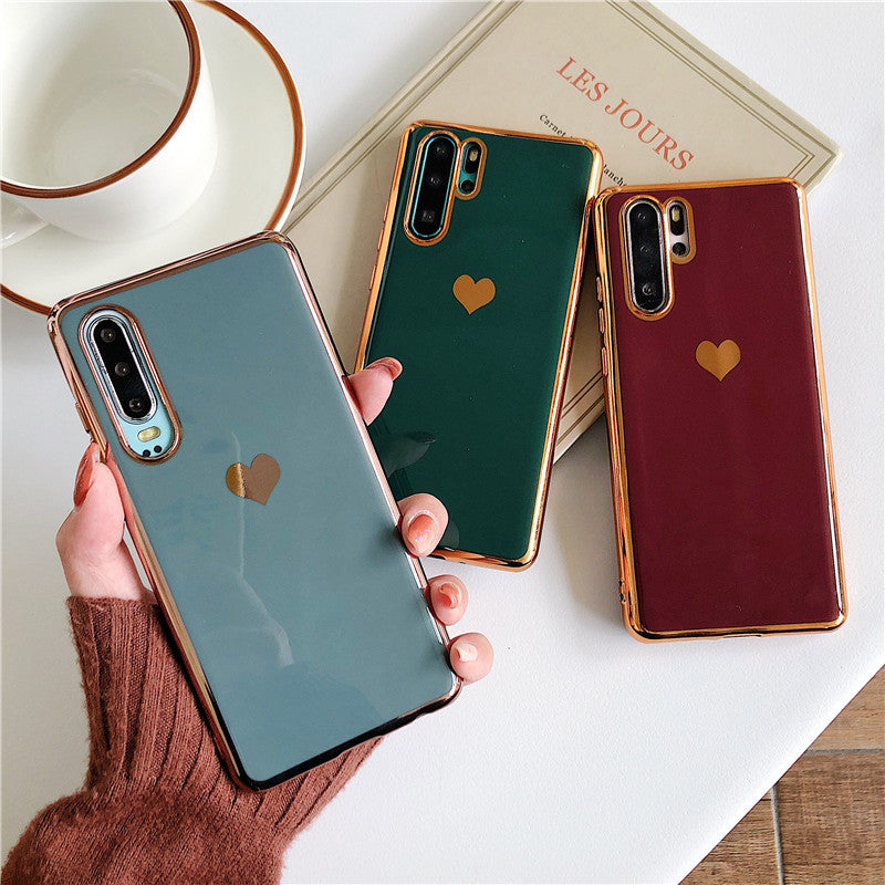Three phone cases with heart designs held by a hand, with a book and cup in the background.