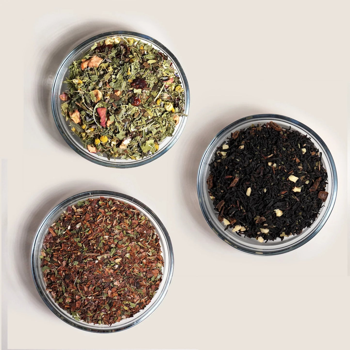 Three glass bowls containing different types of loose leaf tea on a light background